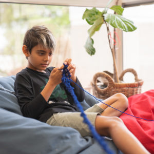a boy sits on a grey chair and knits with blue yarn