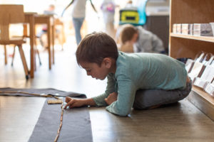 A young boy in a blue shirt kneels on a wooden floor. He is touching a string of gold beads laid out on a gray mat.