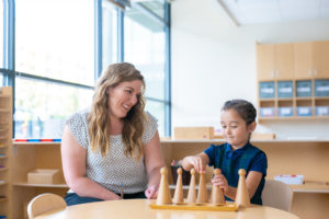 A teacher smiles at a young girl in a blue shirt. They are seated at a wooden table, and the girl is touching a set of wooden cones.