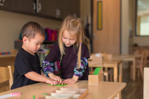 A young girl in a blue shirt leans over to help a younger child with his work. They are both leaning on a wooden table.