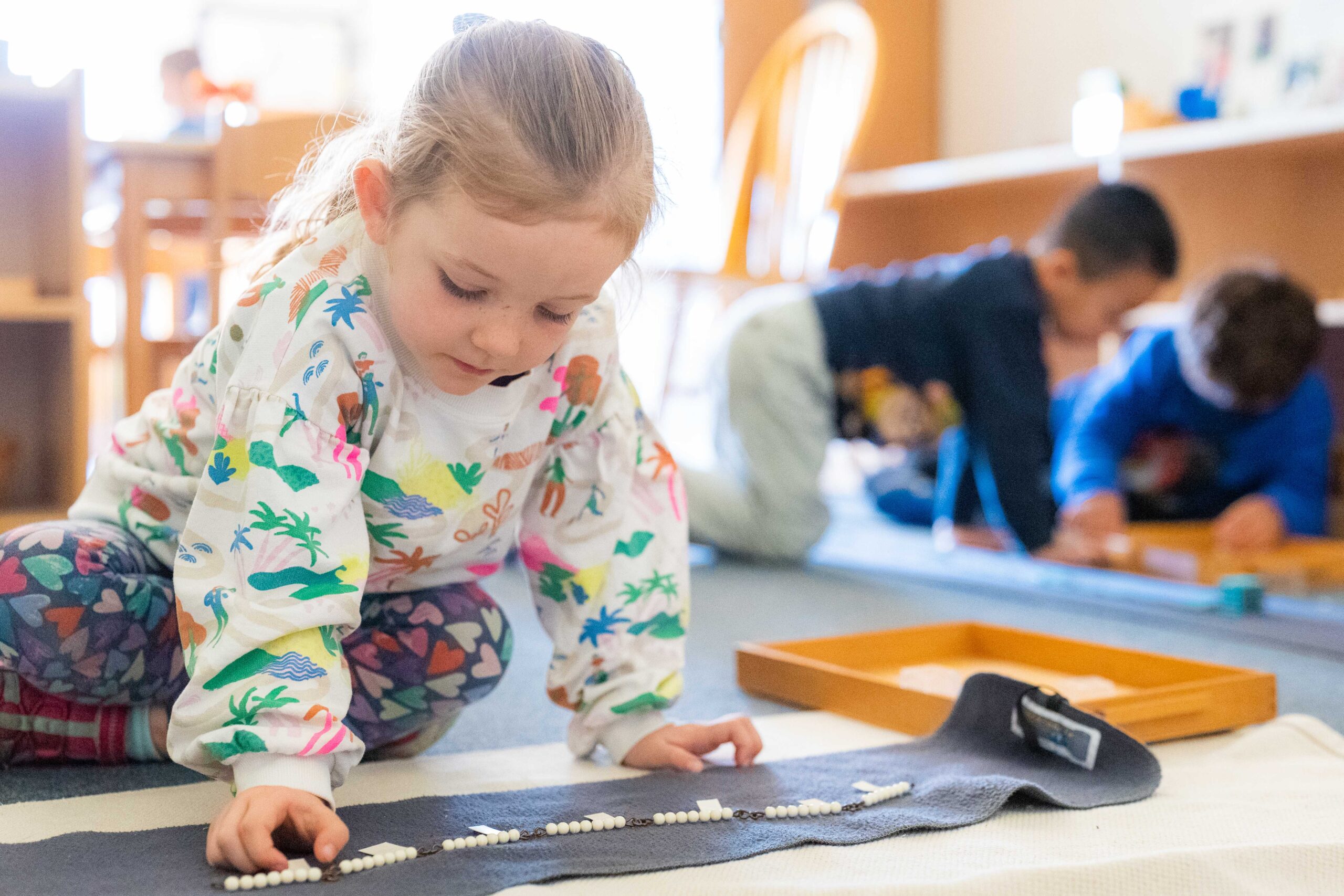 A young girl in a multi-colored sweatshirt moves white beads on a gray rug