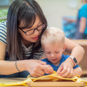 A woman with brown hair and a blue and white stripe top leans over a young child with white hair to help him button