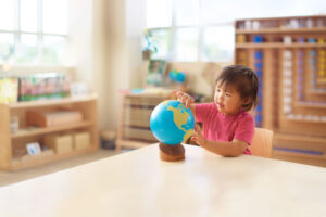 A young girl with black hair and a pink shirt sits at a wooden table and points to a globe