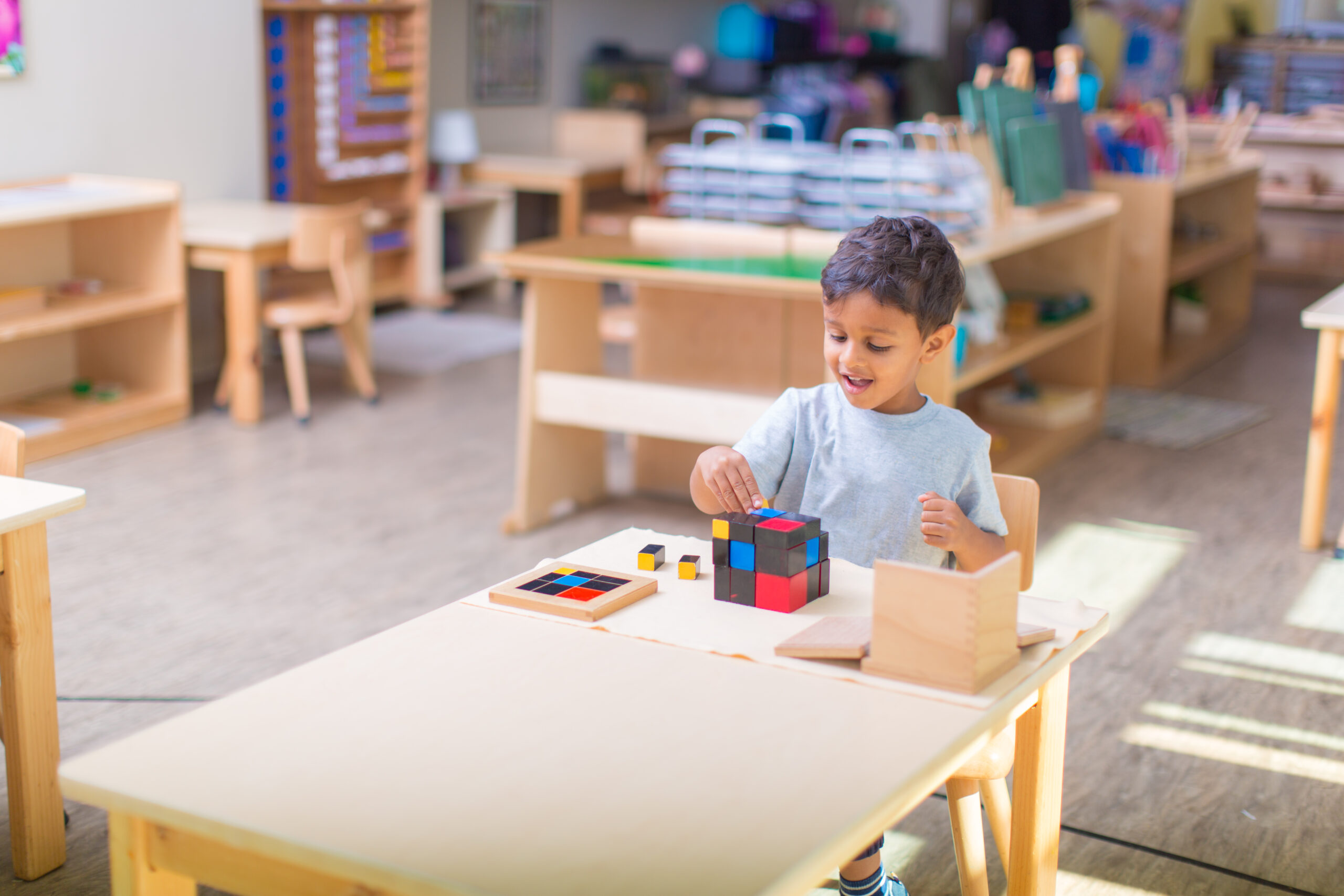 Young boy with dark hair and grey t-shirt sites at a light wood desk and works with a trinomial cube that is colored blue, red, and black