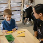 A boy in blue and a teacher in black work with yellow triangles on a wooden table.