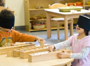 Two children sit at a wooden table. They wear blindfolds and are trying to fit wooden cylinders into the right sized hole in a box.