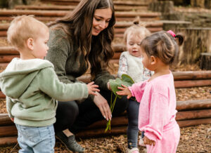 A woman with long brown hair and a green sweater shows a large green leaf to three young children.