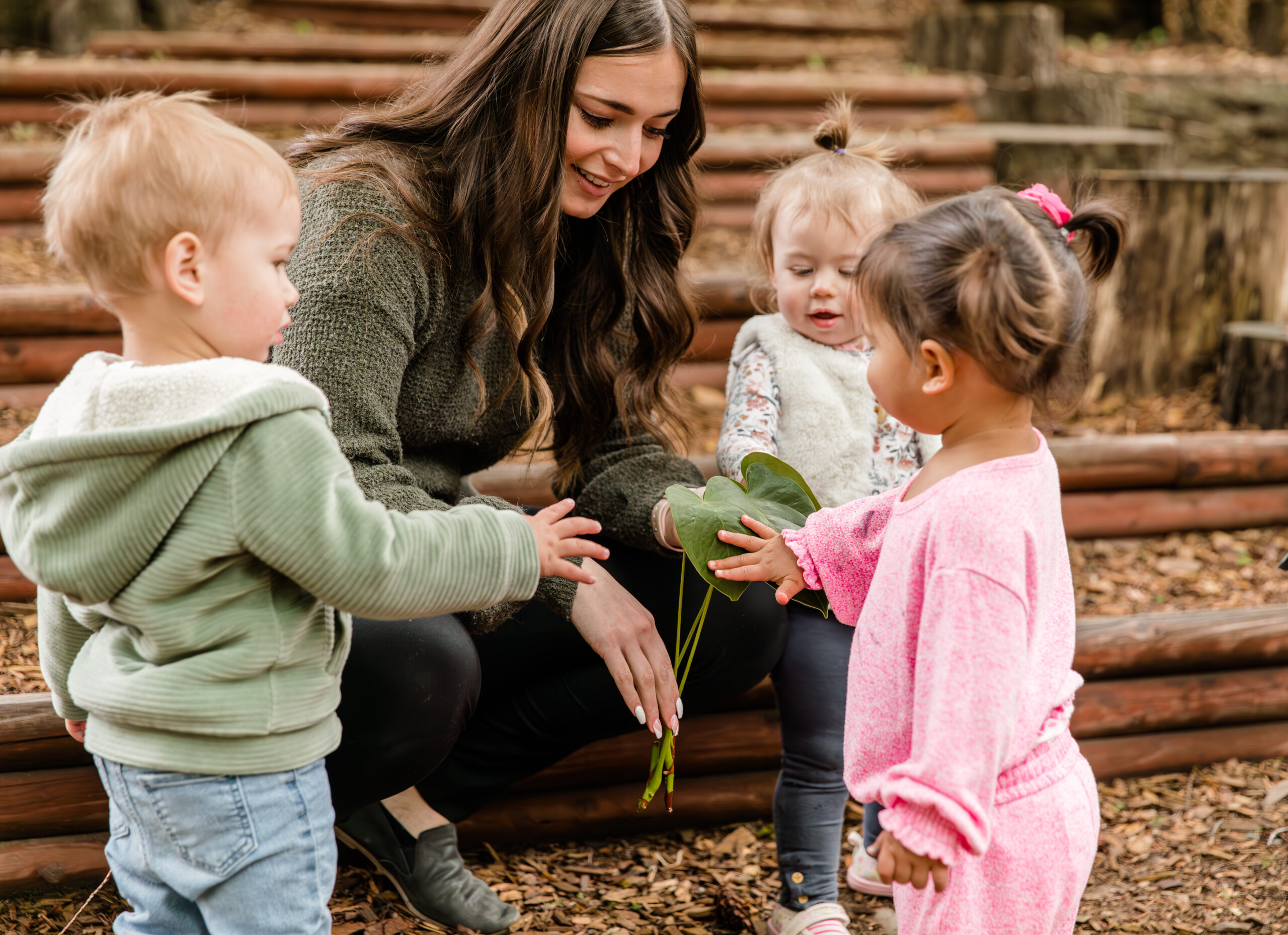 A woman with long brown hair and a green sweater shows a large green leaf to three young children.