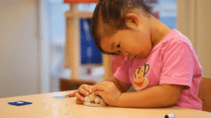 A girl with black hair and a pink shirt carefully threads a needle.